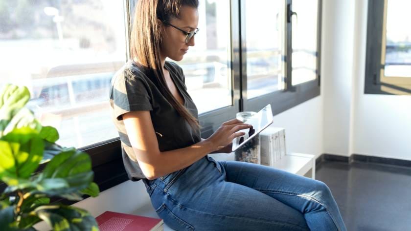 Pretty young business woman using her digital tablet while sitting next to the window in the office.
