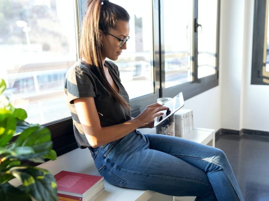 Pretty young business woman using her digital tablet while sitting next to the window in the office.